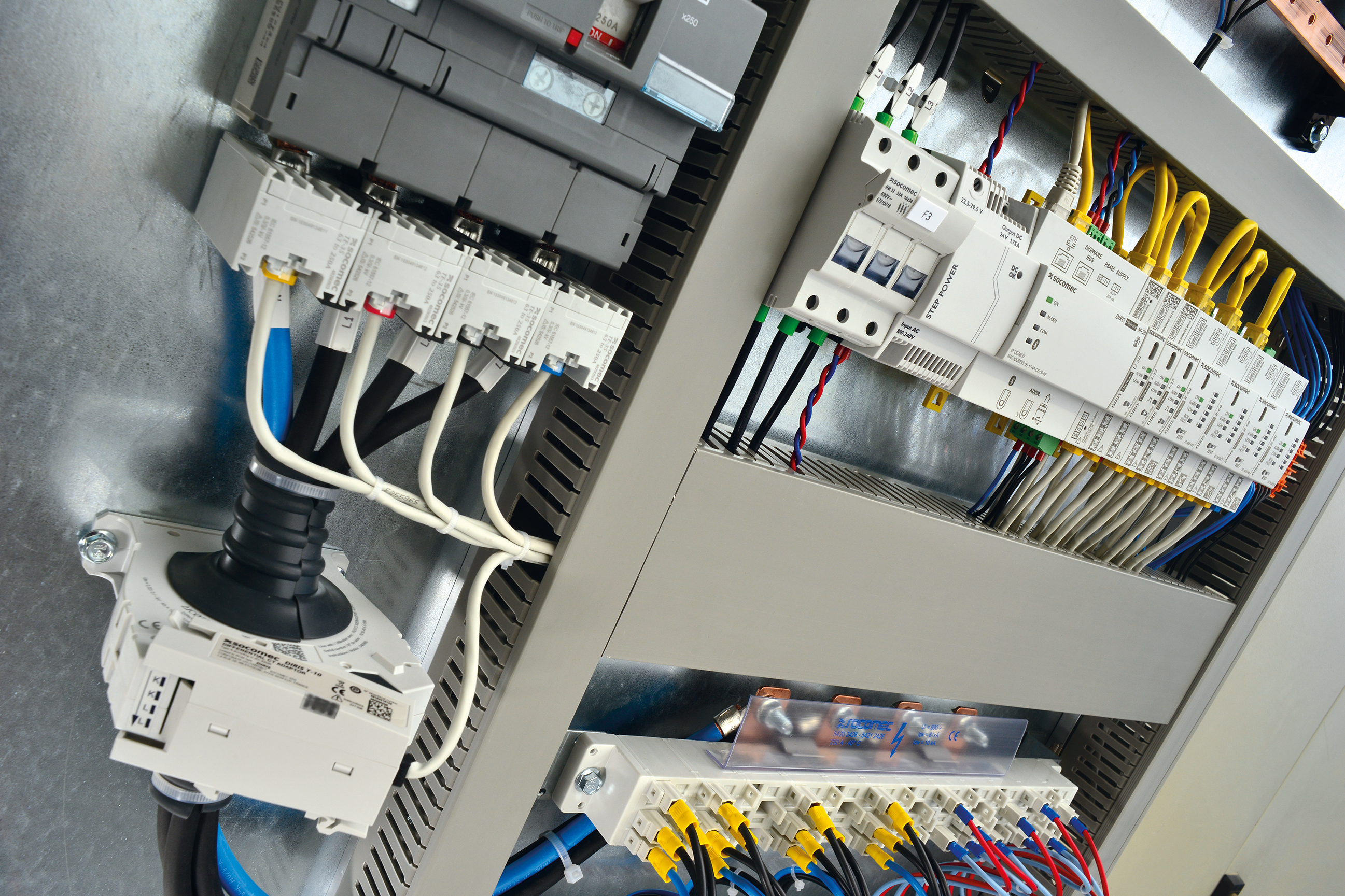Primer plano de un armario eléctrico abierto con componentes SOCOMEC: disyuntores/dispositivos de protección, módulos de control y comunicación, así como bornes y bloques de distribución cableados con conductores de diferentes colores.
